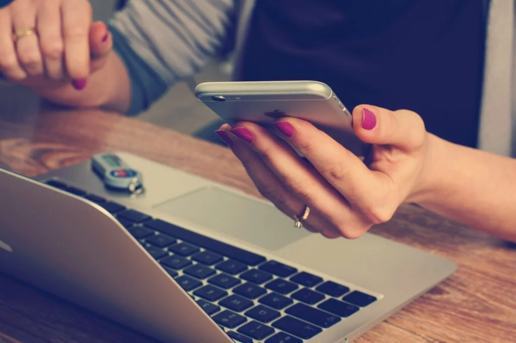 A woman holding a cell phone next to a laptop, possibly considering an upgrade or settling for less.