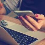 A woman holding a cell phone next to a laptop, possibly considering an upgrade or settling for less.